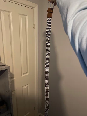 Randigt flätat bälte i blått och vitt - Säljer ett snyggt flätat bälte med randigt mönster i marinblått och vitt. Bältet har detaljer i brunt skinn vid spännet och ett klassiskt silverfärgat spänne. Perfekt accessoar för att lyfta din outfit med lite extra stil.