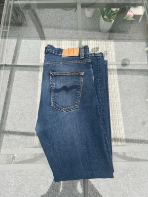 Nudie Jeans Lean Dean New Ink W32 L30 - Säljer ett par Nudie Jeans Lean Dean i den populära tvätten New Ink. Modellen är slim fit i mörkblå denim med lätt stretch, vilket ger en snygg men bekväm passform. Storlek W32 L30.  Jeansen är gjorda i kvalitativ ekologisk denim (99% bomull, 1% elastan) och har de klassiska Nudie-detaljerna med broderade bakfickor och slitstark konstruktion. Jeansen är i fint skick utan fläckar eller skador.  Perfekta om du vill ha stilrena, hållbara och tidlösa herrjeans som funkar till allt.