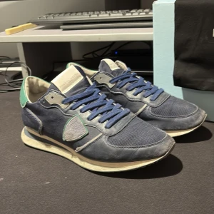 Blå Philippe Model sneakers med gröna detaljer - Snygga sneakers från Philippe Model i marinblått med gröna och vita detaljer. Ovandelen är en mix av skinn och mesh, med blå snörning och rund tå. Platt sula och cool retrostil. Perfekt för dig som gillar streetwear och vill sticka ut med ett franskt märke.