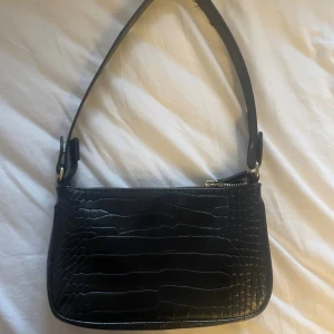 Svart axelväska - Snygg svart axelväska med krokodilmönstrad yta och silverfärgad dragkedja. Väskan har en stilren, rektangulär form och ett brett axelband som ger en cool vibe. Perfekt för att lyfta din outfit med lite edge.