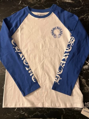 Chrome Hearts long sleeve - Säljer en långärmad topp från Chrome Hearts med vit kropp och blå ärmar. Tryck med Chrome Hearts-logga på bröstet och längs båda ärmarna i blått och vitt. Rund halsringning med blå kant. Materialet är mjuk bomull, perfekt för en cool streetwear-look.