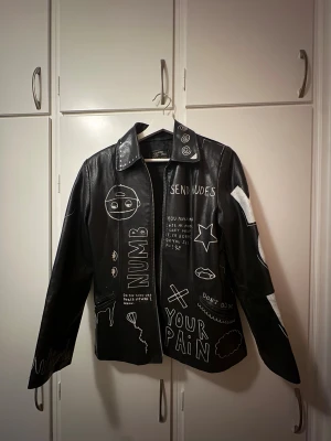 Svart skinnjacka med graffiti-tryck - Unik svart skinnjacka från ett gammalt UF-företag med handmålade vita graffiti-illustrationer och texter över hela jackan. Jackan har klassisk krage, dragkedja framtill och coola detaljer som stjärnor, ögon och olika symboler. Perfekt för dig som vill sticka ut med din stil. Jackan är från Vera Pelle.   Ordinarie pris 700kr