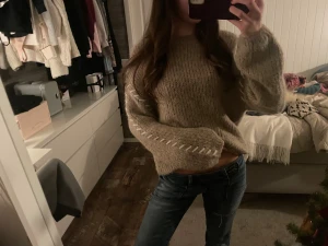 Beige stickad tröja med vita detaljer - Supermysig beige stickad tröja med lång ärm och loose passform. Tröjan har snygga vita snörningsdetaljer längs ärmarna som ger en unik look. Perfekt att slänga på sig när det är kyligt ute. Passar dig som gillar chill och stilren vibe.