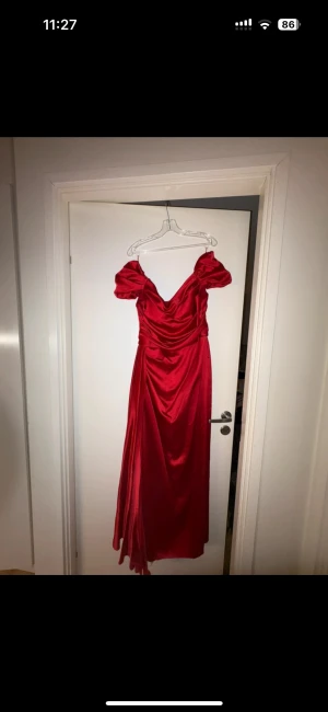 Röd lång satinklänning med slits  - Säljer en röd långklänning i glansigt satinmaterial med offshoulder-ärmar och snygg drapering över byst och höft. Klänningen har en hög slits framtill och en figurnära passform som ger en elegant siluett. Perfekt för bal eller festkväll.