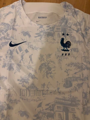 Frankrike Nike fotbollströja mönstrad - Säljer en officiell Frankrike fotbollströja från Nike med unikt ljusblått mönster över hela tröjan. Tröjan är vit med korta ärmar, har FFF-logga med tupp och två stjärnor på bröstet samt Nike Swoosh. Materialet är lätt och andas, perfekt för match eller träning.