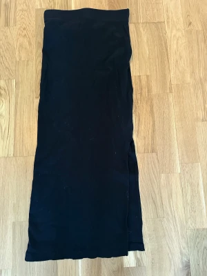 Svart långkjol med slits - Svart långkjol i mjukt tyg med rak passform och en snygg slits på sidan. Kjolen har enkel design utan mönster och resår i midjan för skön komfort. Perfekt för dig som gillar stilrena och minimalistiska plagg.