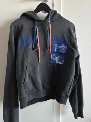 Mörkgrå Off-White hoodie med blått tryck - Säljer en mörkgrå hoodie från Off-White med unikt blått grafiskt tryck fram och bak. Tröjan har huva med snörning i blått och rött, samt en stor magficka. Materialet är mjuk bomull och passformen är normal. Perfekt för dig som gillar streetwear och coola detaljer.
