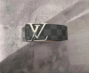 Lv bälte  - Säljer ett Louis Vuitton bälte i svart och grått med klassiskt rutigt Damier-mönster. Bältet har en stor LV-logga i silver på spännet och är tillverkat i äkta skinn. Insidan är ljusbrun med präglad logga och detaljerad söm längs kanterna. Perfekt för dig som gillar lyxiga accessoarer.