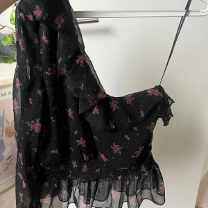 Svart blommig one-shoulder blus med volang - Supersöt svart blus med rosa blommor och volangdetaljer. Blusen har omlott-design, oneshoulder och smockad midja som ger en snygg passform. Tunn och luftig polyester. Blommigt mönster och transparent nederkant för extra vibe. Helt oanvänd med ordinariepris på 249kr