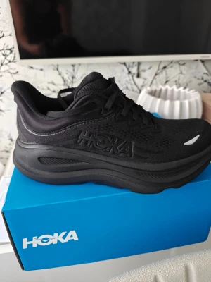 Svarta Hoka sneakers med tjock sula - Säljer ett par helsvarta Hoka sneakers med riktigt chunky sula och snyggt broderad logga på sidan. Skorna har snörning och är tillverkade i mesh och syntetmaterial för extra komfort. Perfekta för dig som gillar sportig och modern stil.