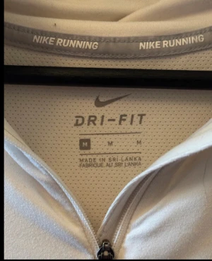 Vit Nike långärmad träningströja - Nike Dri-Fit långärmad träningströja i vit med gråa ränder på ärmarna. Tröjan har half zip framtill och ett reflekterande Nike-logga på bröstet. Perfekt för löpning eller gym, med lätt och ventilerande polyester-material.