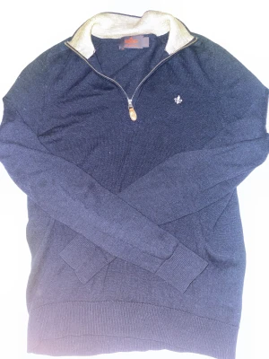 Marinblå half zip tröja Morris S - Snygg marinblå stickad tröja från Morris i 100% merinoull. Tröjan har en beige krage, half zip-dragkedja och en broderad vit logga på bröstet. Perfekt för dig som gillar klassisk stil med en modern touch.