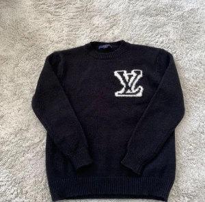 Svart stickad tröja Louis Vuitton L - Svart stickad tröja från Louis Vuitton i storlek L. Tröjan har ett stort vitt LV-logo broderat på bröstet och ribbade muddar vid ärmslut och nederkant. Perfekt för dig som gillar exklusiva märken och stilren design.