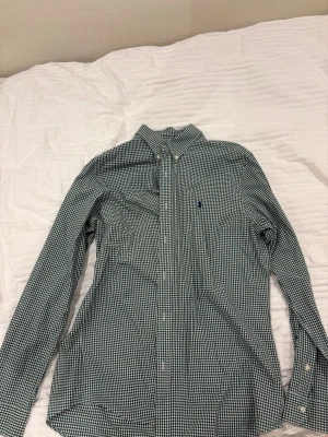 Rutig grön skjorta från Polo Ralph Lauren - Snygg rutig skjorta från Polo Ralph Lauren i grönt och vitt, custom fit. Skjortan har klassisk krage med knappar, lång ärm och broderad logga på bröstet. Tillverkad i mjuk bomull med lite stretch för extra komfort. Perfekt för dig som gillar stilren och tidlös design.
