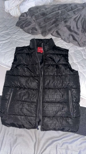 Svart dunväst från Guess - Svart äkta dunväst från Guess med hög krage och två dragkedjefickor. Västen har ett diskret mönster med Guess-loggor över hela ytan och en Guess-triangel på bröstet. Materialet är glansigt och quiltat, perfekt för att hålla värmen under kyliga dagar.