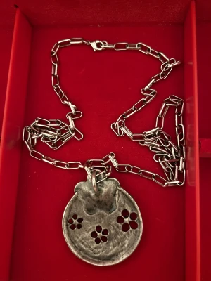 Silvrigt halsband med blommor - Unikt halsband i silverfärgad metall med grov kedja och stor rund berlock dekorerad med blommor och små stenar. Berlocken har en hamrad yta och utstansade blommor, samt en upphöjd blomma i mitten. Perfekt för dig som gillar statement-smycken.