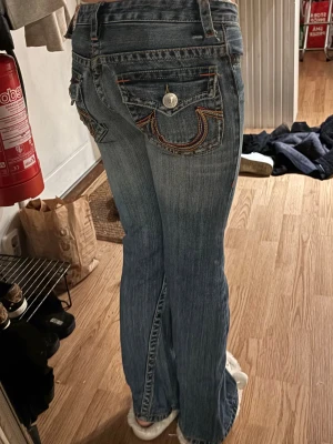 True religion jeans  - Säljer ett par blå jeans från True Religion med låg midja och bootcut passform. Jeansen har snygga broderade bakfickor med silverfärgade knappar och kontrastsömmar. Klassisk jeanslook med slitningar och detaljer som sticker ut.