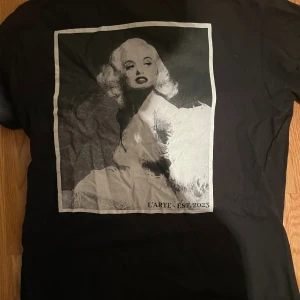 Svart t-shirt  - Svart t-shirt från L'ARTE med stort grafiskt konstmotiv i gråskala på ryggen och texten 'L'ARTE EST. 2023'. Klassisk passform och rund hals. Perfekt för dig som gillar streetstyle och unika prints. Skriv vid funderingar🤝