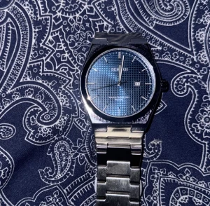 Blå DENISSI klocka med stållänk - Stilren klocka från DENISSI med blå urtavla och diskret rutmönster. Klockan har en robust silverfärgad länk i metall och datumvisning. Klassisk rund boett och minimalistiska indexmarkeringar ger en modern look.