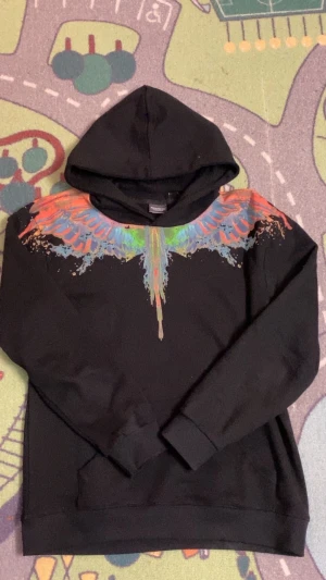Svart hoodie Marcelo Burlon med färgglada vingar - Svart hoodie från Marcelo Burlon County of Milan med färggrafiskt vingtryck i orange, blått, grönt och rosa över axlar och bröst. Tröjan har huva, långa ärmar och ribbade muddar. Perfekt för dig som gillar streetwear med unik design.