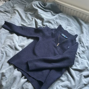 Mörkblå stickad tröja Polo Ralph Lauren - Mörkblå stickad tröja från Polo Ralph Lauren med halv dragkedja i halsen. Tröjan har lång ärm och en klassisk, lite högre krage. Materialet är mjukt och värmande, perfekt för kyliga dagar. Snygg och enkel design med diskret logga på bröstet. Tröjan är knopprig 