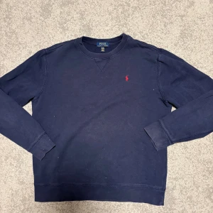 Mörkblå sweatshirt från Polo Ralph Lauren - Snygg mörkblå sweatshirt från Polo Ralph Lauren i storlek XL. Tröjan har lång ärm och klassisk rund hals. På bröstet finns det ikoniska röda Polo-broderiet. Perfekt för dig som gillar stilrena och tidlösa plagg.