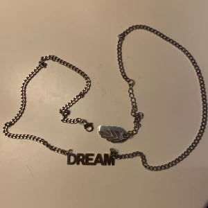 Silverfärgat halsband med DREAM-text - Snyggt halsband i silverfärg med en kedja och ett hänge där det står DREAM i stora bokstäver. På kedjan finns även en liten platta formad som ett löv. Perfekt för dig som gillar statement-smycken med personlig touch.