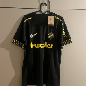 AIK svart matchtröja Nike M - AIK:s officiella svarta fotbollströja från Nike i storlek M. Tröjan har gula detaljer, klubbmärke på bröstet, sponsortryck och mönstrade muddar. Materialet är lätt och andas, perfekt för träning eller match. Snygg design med tryck både fram och bak.