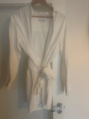 Vit skjorta/ klänning  - Säljer en stilren vit skjorta/ klänning med lång ärm och knyt i midjan. Klänningen har klassisk krage och är tillverkad i ett lätt bomullstyg, perfekt för en fräsch och enkel look.