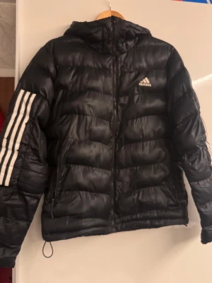 Svart Adidas pufferjacka med huva - Svart pufferjacka från Adidas med huva och klassiska vita ränder på ärmarna. Jackan har dragkedja framtill och Adidas-logga på bröstet. Tillverkad i syntetmaterial och har en quiltad design som ger extra volym och värme. Perfekt för kalla dagar.