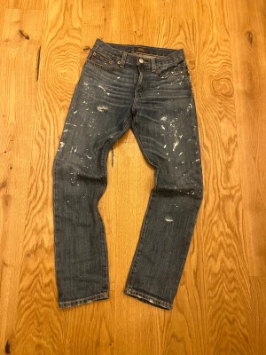 Mörkblå Polo Ralph Lauren jeans - Säljer ett par mörkblå jeans från Polo Ralph Lauren med coola slitningar och vita färgstänk. Klassisk femficksmodell, rak passform och normal midja. Jeansen är i robust denim och har snygga detaljer som synliga sömmar och läderpatch bak.