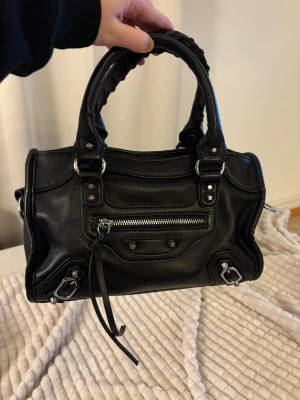 Svart handväska med silverdetaljer - Cool svart handväska i skinn med snygga silverdetaljer och dragkedjor både fram och bak. Väskan har två robusta handtag med flätad look och röd insida med flera fack. Perfekt storlek för det viktigaste och ger en edgy vibe till din outfit. Köpt för 1000kr, säljer för 800!