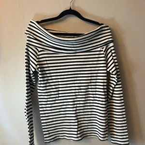 Randig offshoulder topp i bomull - Säljer en vit och svart randig topp med offshoulder-design och långa ärmar. Toppen har en bred krage som vilar snyggt över axlarna och är gjord i mjuk bomull. Perfekt för dig som gillar en stilren och chill look. Från Cubus i strl L. 