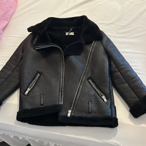 Svart skinnjacka från NA-KD - Snygg svart skinnjacka från NA-KD med fuskpälsfoder och asymmetrisk dragkedja. Jackan har långa ärmar och flera dragkedjedetaljer. Perfekt för kyligare dagar.