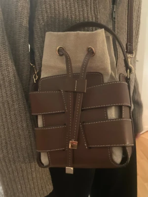 Bucket bag HM - Säljer en snygg axelväska från HM med cool design i brunt skinn och beige canvas. Väskan har ett unikt flätat mönster med snörning framtill, guldfärgade detaljer och både handtag och axelrem. Perfekt storlek för det viktigaste och ger en trendig vibe.