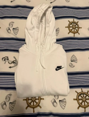Vit hoodie från Nike med dragkedja - Vit hoodie från Nike med svart logga på bröstet. Tröjan har huva med snörning, två fickor fram och hel dragkedja. Tillverkad i mjuk bomullsmix med fleece på insidan för extra komfort. Perfekt för chill eller träning.