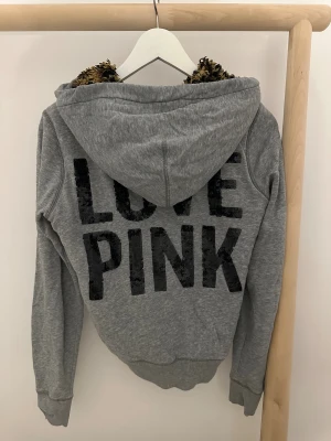 Grå hoodie PINK med paljetter XS - Grå hoodie från PINK by Victoria's Secret i storlek XS. Utsidan har svart paljetttext 'LOVE PINK' på ryggen och  är fodrad med fluffigt leopardmönstrat tyg. Dragkedja framtill, snörning vid huvan och känguruficka. 