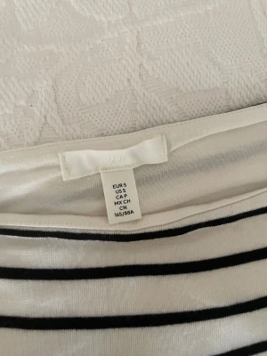 Randig långärmad body från H&M - Säljer en snygg långärmad body från H&M i storlek S. Den är vit med svarta horisontella ränder och har en rundad hals. Materialet är mjukt och stretchigt, troligen bomull med inslag av syntet. Perfekt att styla med jeans eller kjol för en cool look.