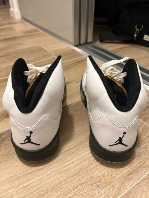 Nike Air Jordan 5 vit/guld - Säljer ett par Nike Air Jordan 5 sneakers i vitt skinn med svarta detaljer och guld på plösen. Klassisk Jumpman-logga på hälen, snörning framtill och svart sula. Grymma high-top sneakers med ikonisk design och coola guldaccenter.