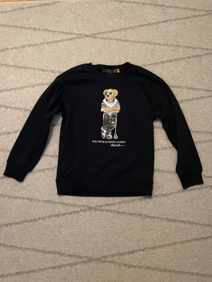 Svart sweatshirt Polo Bear Ralph Lauren - Svart sweatshirt från Polo Ralph Lauren i storlek S med ikoniskt Polo Bear-tryck på bröstet. Tröjan har rund hals, ribbade muddar och är långärmad. Materialet är mjuk bomull, perfekt för en chill och stilren look.