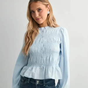 Ljusblå blus med volang och knappar - Supersöt ljusblå blus med volangkant nedtill och smockad front. Blusen har långa puffärmar och knappar hela vägen framtill. Perfekt att styla med jeans eller kjol för en fräsch look. Materialet känns lätt och luftigt.