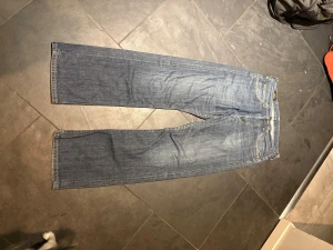 Blå Lee jeans med raka ben - Säljer ett par klassiska blå jeans från Lee med raka ben och normal passform. Jeansen har fem fickor, gul kontrastsöm och Lee-lapp i brunt läder baktill. Materialet är slitstarkt denim i bomull och färgen är mellanmörk blå med lätt tvättad look. De har en rak-loose passform. Storleken är 31/32. De är sparsamt använda.