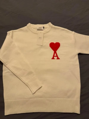 Vit stickad tröja från Ami Paris - Vit stickad tröja från Ami Paris med rund hals och ribbade muddar. På bröstet finns ett stort rött hjärta och bokstaven A som signaturdetalj. Tröjan har lång ärm och är gjord i mjukt material som känns skönt mot huden. Perfekt för dig som gillar stilrena plagg med en twist.