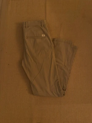 Beige chinos från Jack & Jones - Säljer ett par stilrena beige chinos från Jack & Jones, modell Slim/Marco. Byxorna har klassiska fickor fram och bak, bälteshällor och knappstängning. Perfekta för en clean och avslappnad look. Materialet är mjukt och skönt bomullstyg.