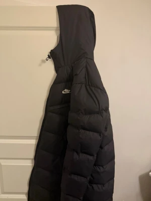 Svart Parka pufferjacka från Nike - Säljer en svart lång pufferjacka från Nike med huva och dragkedja. Jackan har en quiltad design och är tillverkad i syntetmaterial. Perfekt för kalla dagar och har en clean look med Nike-logga på bröstet.