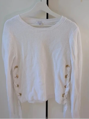 Vit stickad tröja med snörning från Amisu - Säljer en vit stickad tröja från Amisu i storlek M. Tröjan har snygga snörningsdetaljer med guldfärgade öljetter på båda sidorna och är ribbstickad för en modern look. Långärmad och perfekt för lager på lager under höst och vinter.