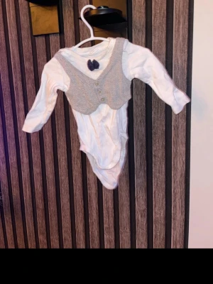 Grå body med väst till baby storlek 62 - Säljer en söt grå body med väst,  med v-ringad form och tre knappar framtill. Bodyn är i ett mjukt stickat material och har ett diskret mönster. Perfekt att styla för en gullig look. Storlek 62