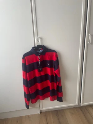 Randig rugbytröja från Gant röd/svart - Säljer en klassisk rugbytröja från Gant i storlek M. Tröjan har breda röda och svarta ränder, krage i svart och Gant-logga broderad på bröstet. Tillverkad i mjuk bomull som känns skön mot huden. Perfekt för dig som gillar sportig och tidlös stil.
