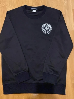 Svart Chrome Hearts sweatshirt - Svart sweatshirt från Chrome Hearts med vit logga på bröstet och stor Chrome Hearts New York-tryck på ryggen. Tröjan har rund hals, ribbade muddar och är långärmad. Perfekt för dig som gillar streetwear och vill sticka ut med en ikonisk design.