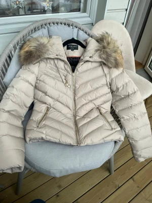 Beige jacka från Hollies stl 38 - Säljer en beige jacka från Hollies i mycket fint skick. Jackan har flera dragkedjefickor, snygga guldfärgade detaljer och quiltad design. Storlek 38 men liten i storlek så passar en 36 också.